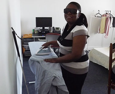 A woman ironing a shirt. She is wearing dark glasses and a white cane leans against the wall nearby.