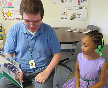 A young man sitting in a chair reading a book to a little girl, who is also sitting in a chair.