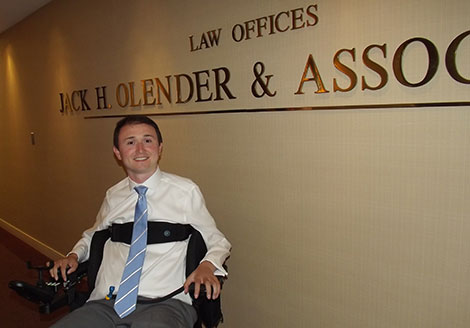 Josh sitting in the hallway in front of his employer's sign: Law Offices, Jack H. Olender & Associates.
