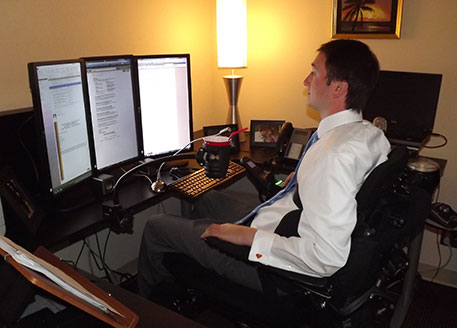 Josh at his desk with assorted assistive technology, including: three legal-size computer screens and a drinking cup holder.