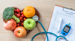 A bowl of fruit and vegetables,a diabetes meter, and a clipboard, on a wooden surface.