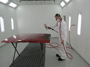 A man in a respirator and white coveralls spray paints a car hood in a body shop paint booth.