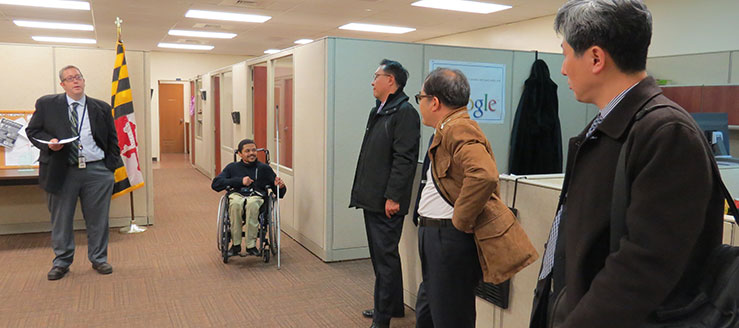 Five men in a office area. One man talks and the others lister. The man in the center is in a wheelchair and holds a white cane.