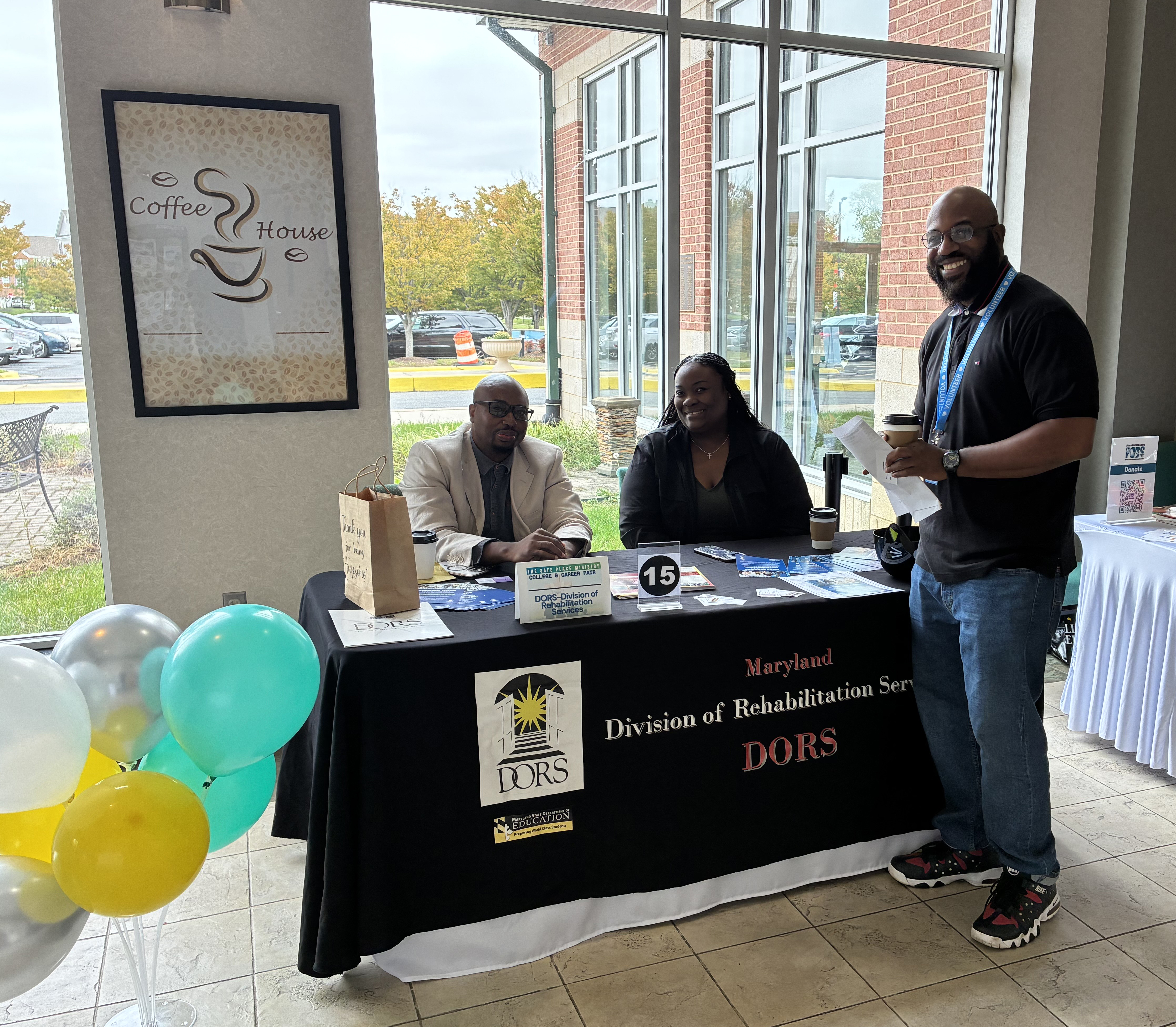 A mand and a women sitting at DORS table at a resource fair, and a man standing in front of the table.