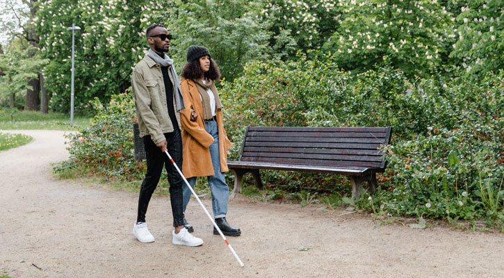 A man and woman walking in a park. He carries a white cane and is holding the woman's arm.