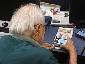An elderly man using a handheld video magnifier to read the newspaper.