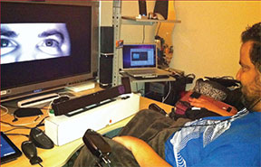 A man in a motorized chair sits in front of an array of computer equipment, including a large flat screen monitor on which is displayed a close-up of his eyes.