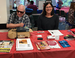 A man wearing dark glasses and a woman, sitting at a table, at an outreach event. The table has several DORS publications on it.