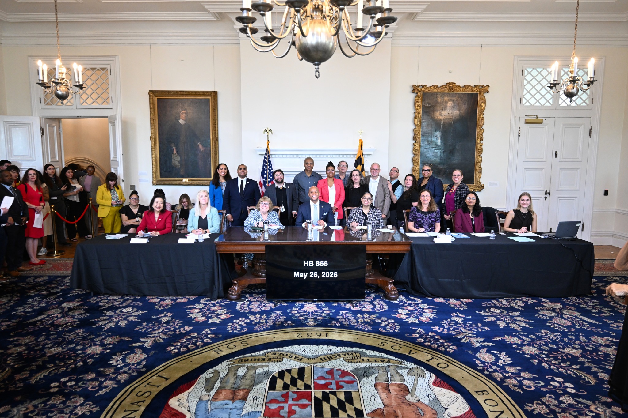 A group of approximately 30 people standing behind, or sitting at a table in the Governor's Reception Room, while Governor Wes M