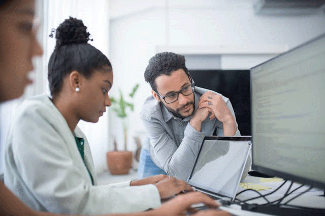 Two women sitting at computer screens with their hands on keyboards. A standing man is leaning in to look at one of the screens.