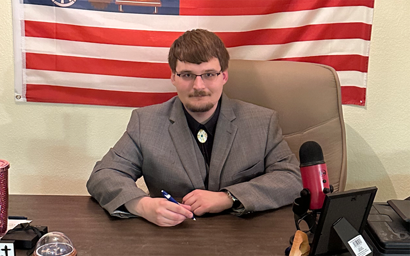 Ben, sitting at a desk, wearing a suite and holding a pen. There is a red and white striped flag hanging behind him.