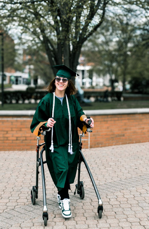 Lily using a standing walker on the campus of Wright State University in a green graduation gown with white tassels.