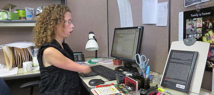 A young woman sitting in an office cubicle, looking at a computer screen. Her right arm ends at the elbow.