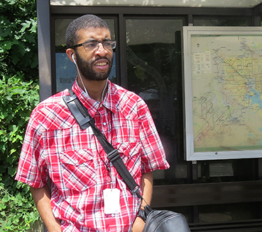 A young man standing at a bus stop with a bus route map behind him.